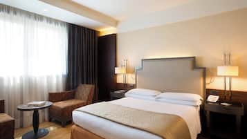 A hotel room with a large bed, two bedside tables with lamps, a small round table with a book, a sofa, and a window with curtains.