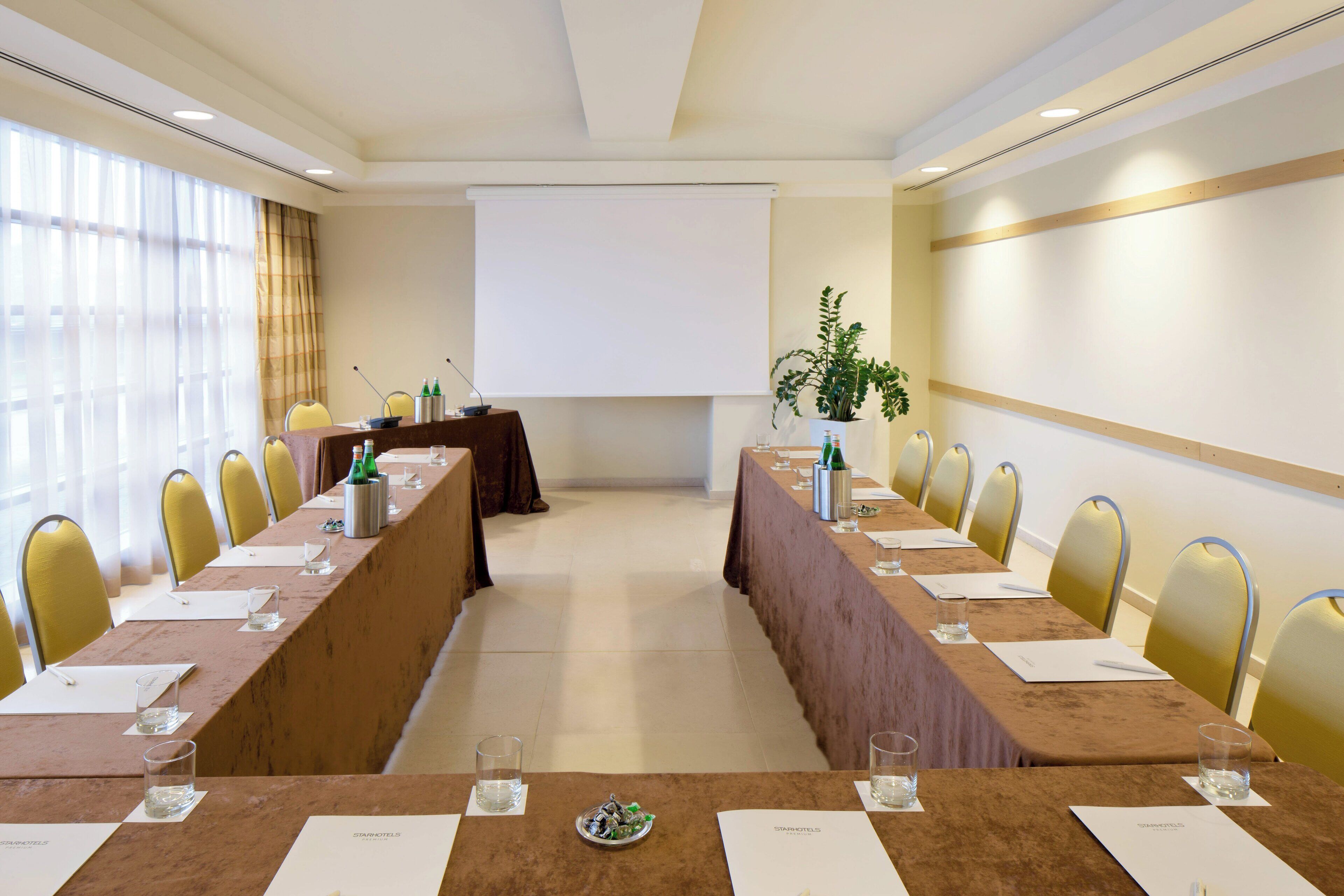 Un salón de conferencias con una larga mesa, sillas, una pantalla de proyector y una planta en maceta.