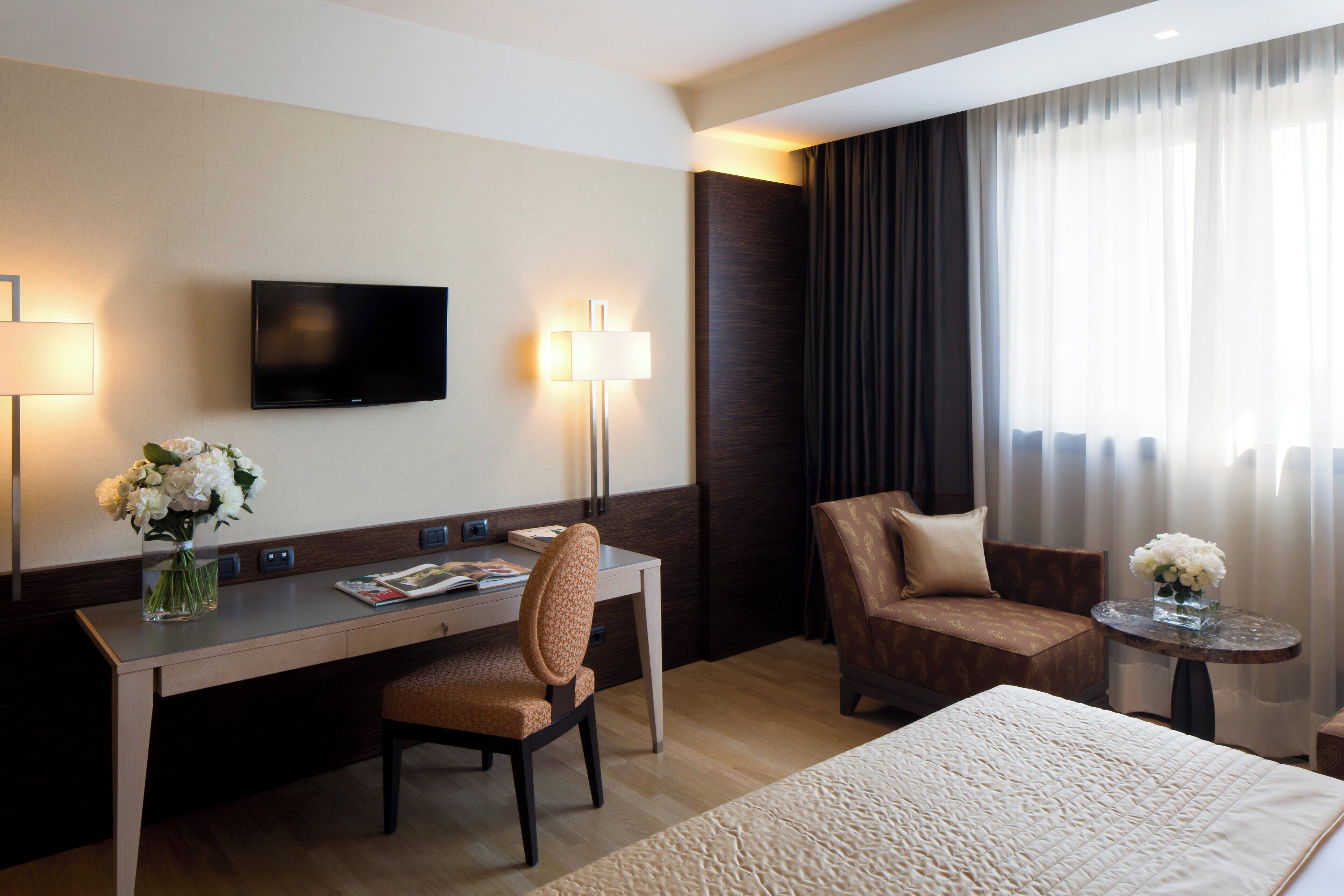 Ein Hotelzimmer mit einem Bett, einem Schreibtisch mit Stuhl, einem an der Wand montierten Fernseher und einem kleinen Tisch mit Blumen.