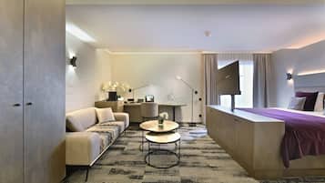 A modern hotel room with a sofa, a round table, a bed with a purple blanket, and a desk with a lamp.