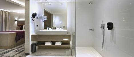 Un baño de hotel moderno con ducha envuelta en vidrio, tocador con espejo, y una cama en la habitación contigua.