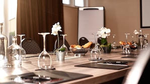 A conference table set with glassware, flowers, and a bowl of pastries in a modern room with a projector screen in the background. - Van Der Valk Hotel Charleroi Airport (Charleroi)