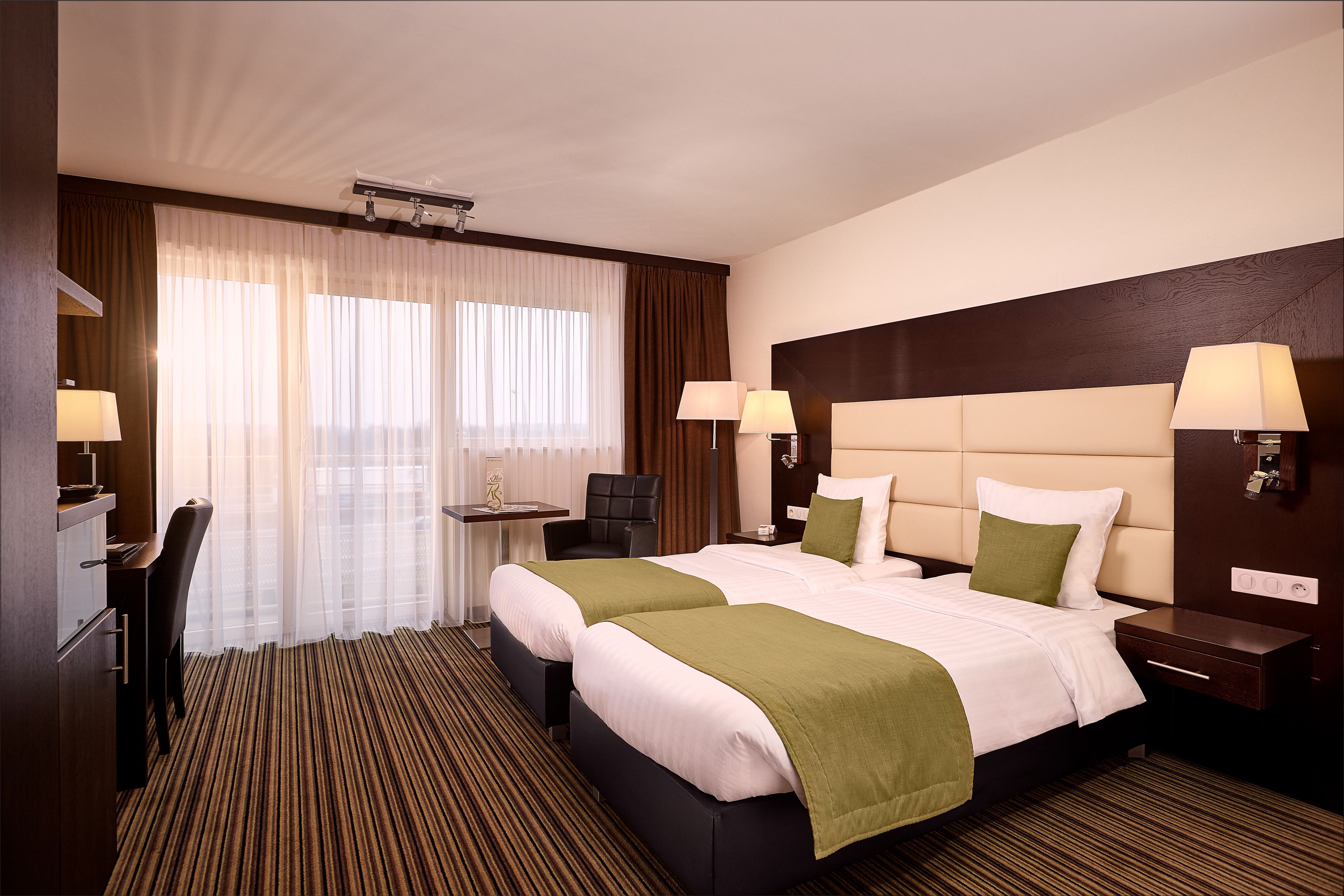 A hotel room with two beds, a desk, a chair, and a large window with curtains.