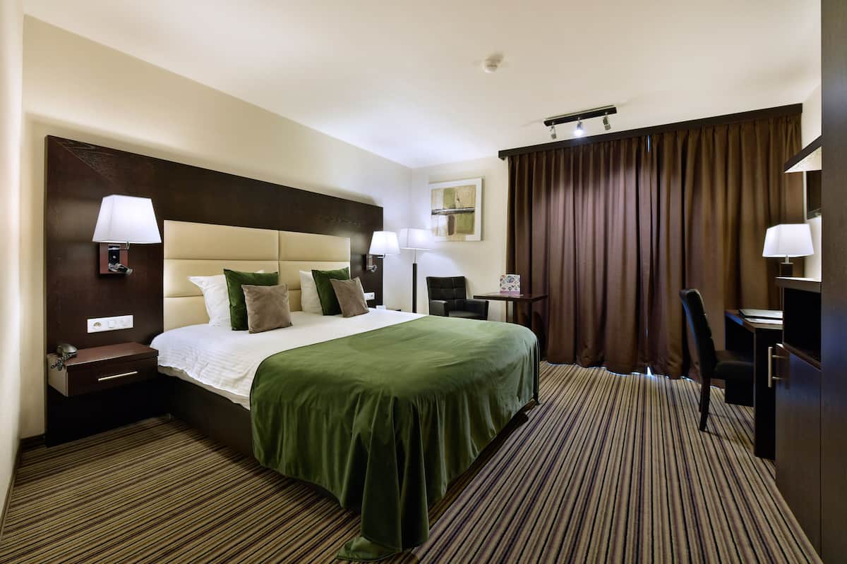 Ein Hotelzimmer mit einem großen Bett, zwei Nachttischen, einem Schreibtisch, einem Stuhl und einem großen Fenster mit Vorhängen.