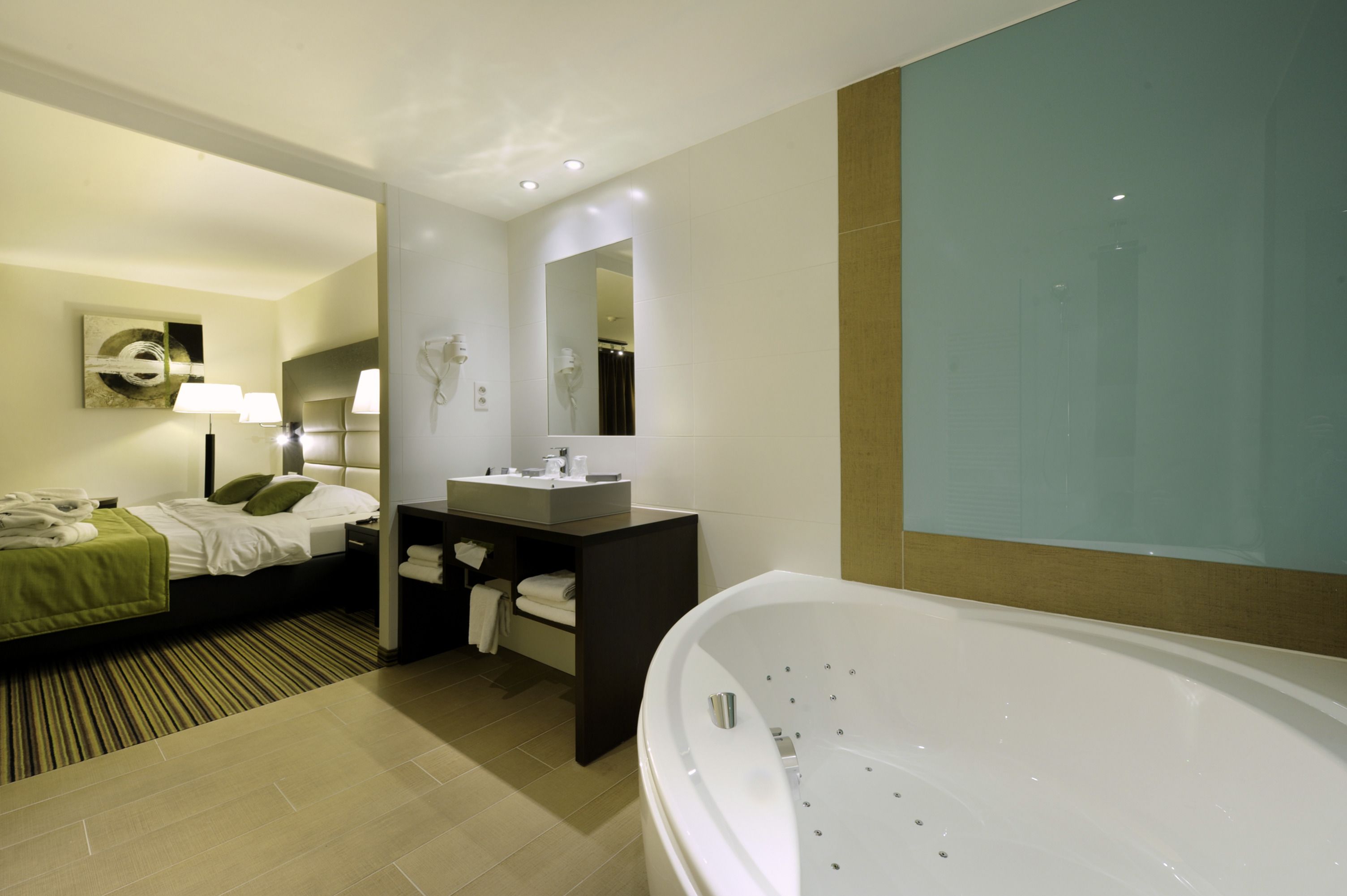 Une salle de bain d’hôtel moderne avec une grande baignoire, un lit avec une literie verte, une coiffeuse avec un miroir et un porte-serviettes.