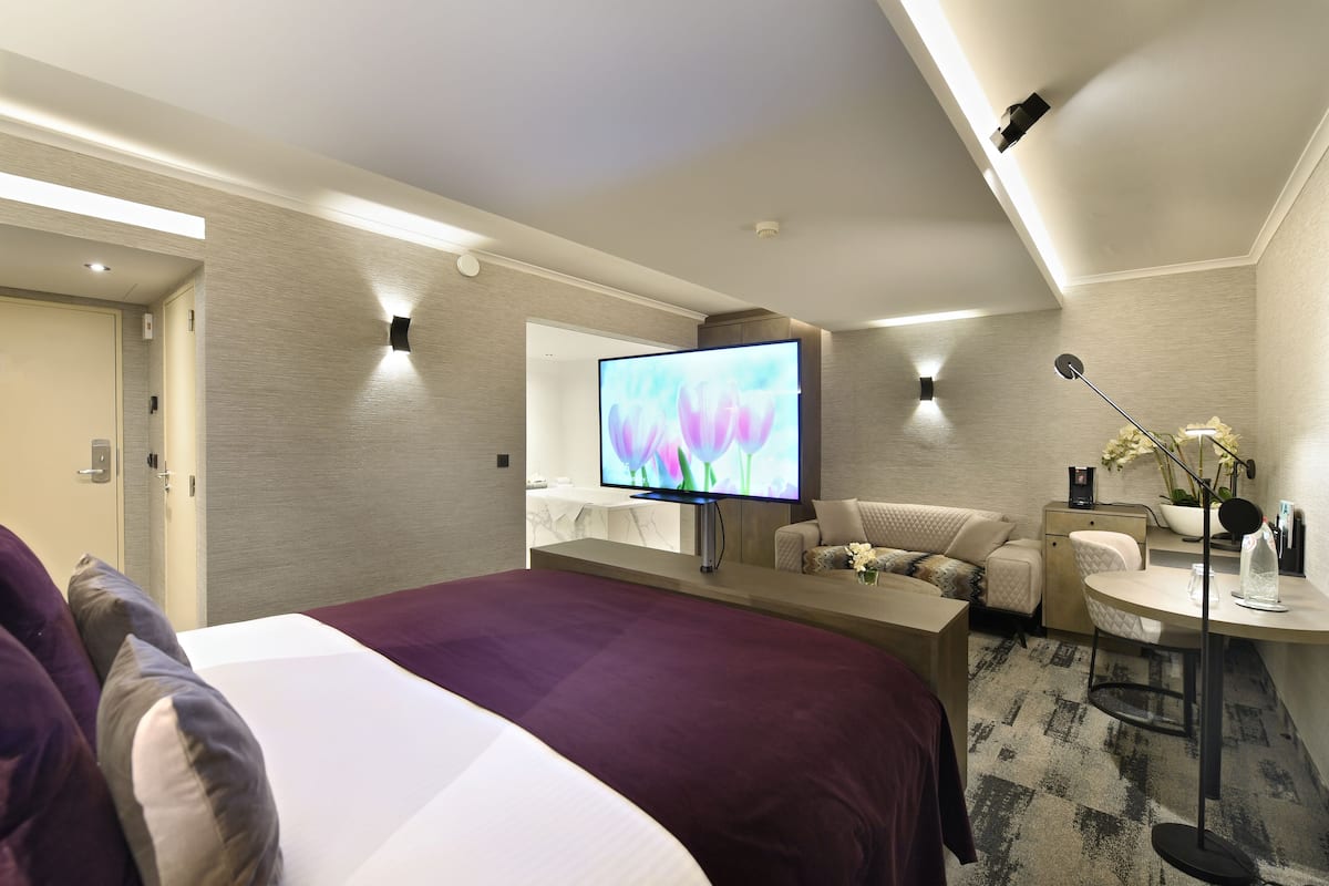 Ein modernes Hotelzimmer mit einem großen Bett, einem Schreibtisch mit Stuhl, einem Fernseher und einem kleinen Tisch mit Lampe.