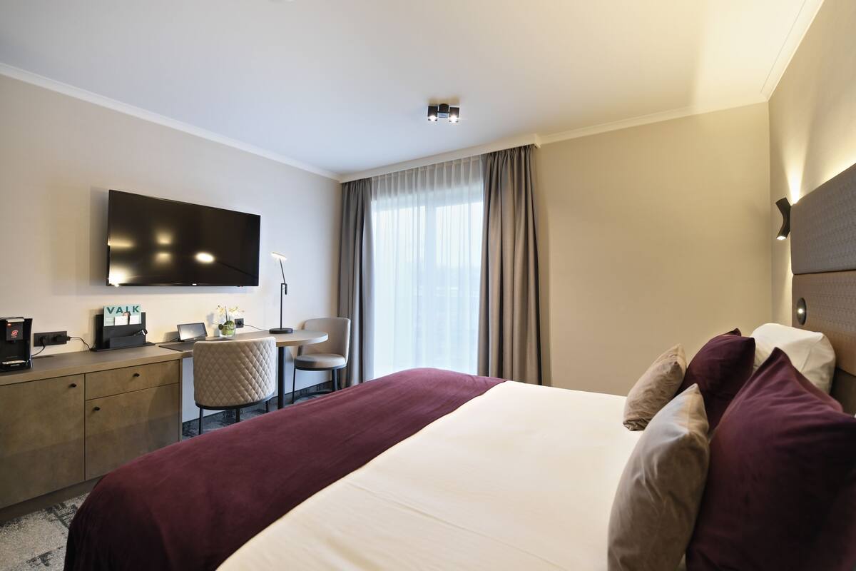 Ein Hotelzimmer mit einem Bett, einem Schreibtisch, einem Sessel, einem Fernseher und einem fensterbelaubten Fenster.