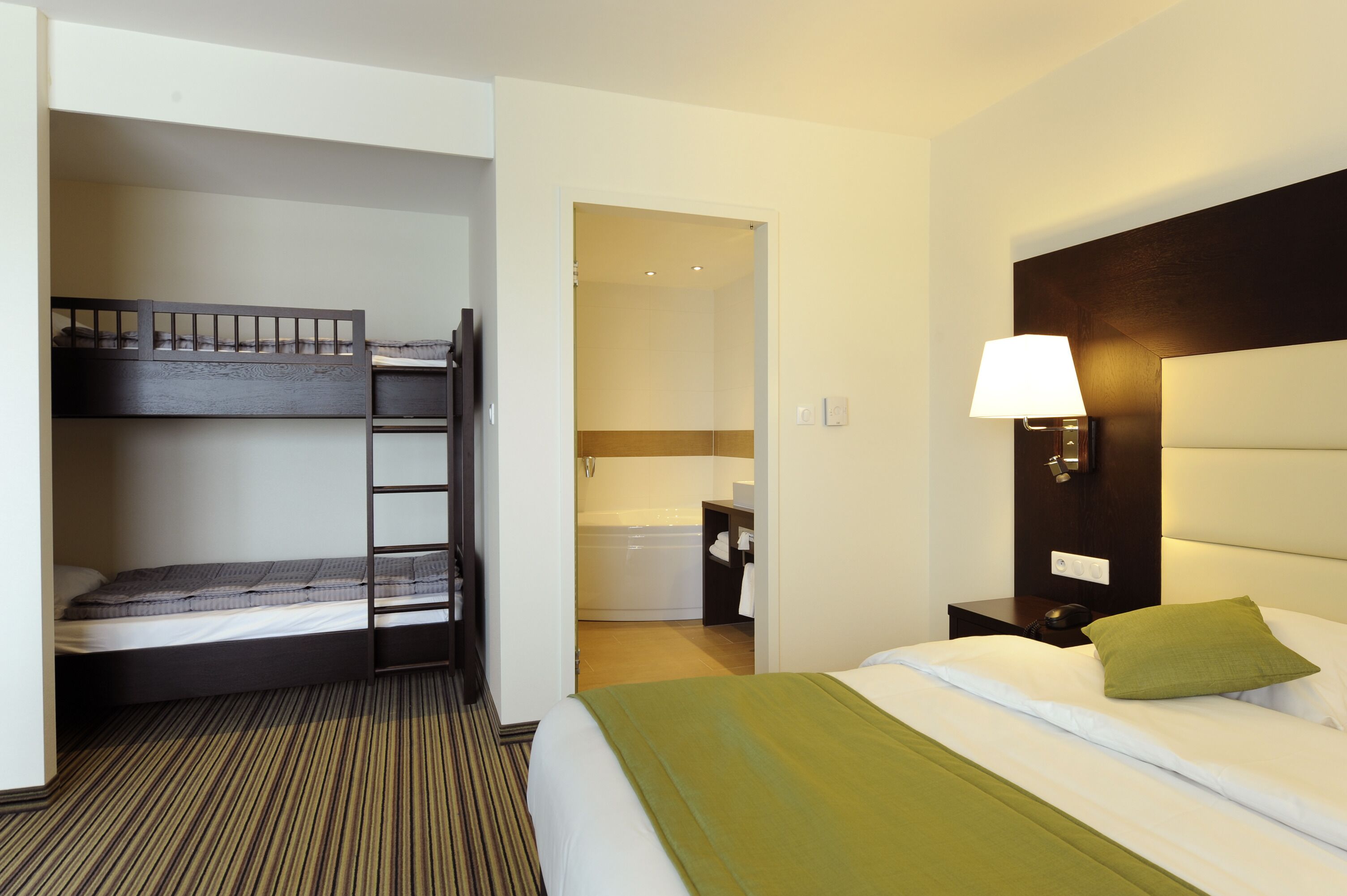 Habitación de hotel con literas, una cama individual, baño con bañera y una mesita de noche con lámpara.