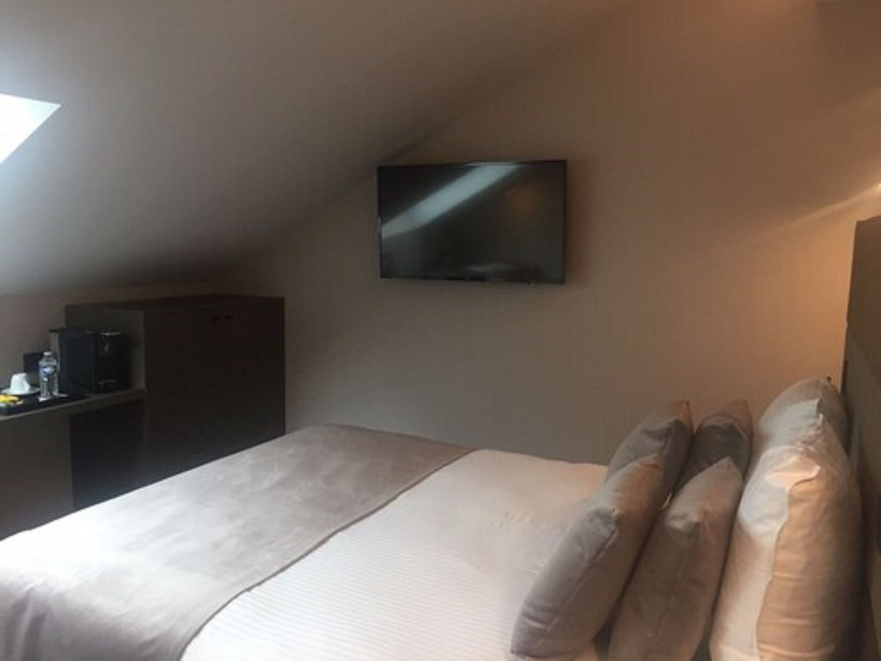 A hotel room with a bed, pillows, a TV mounted on the wall, a desk with a coffee maker, and a wardrobe.