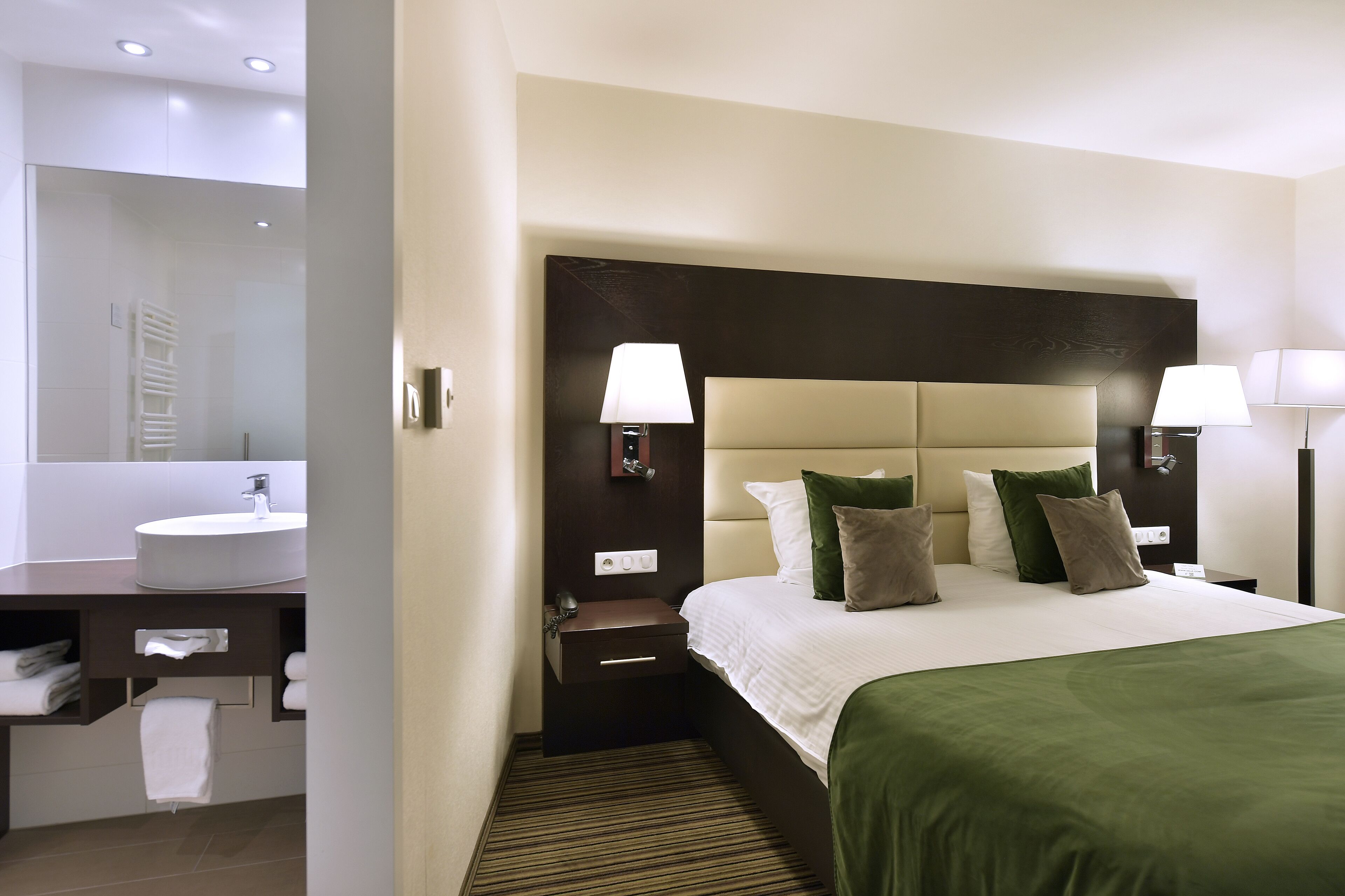 Une chambre d’hôtel avec un grand lit, deux tables de chevet avec des lampes, une salle de bain avec un lavabo et un miroir, et un porte-serviettes.