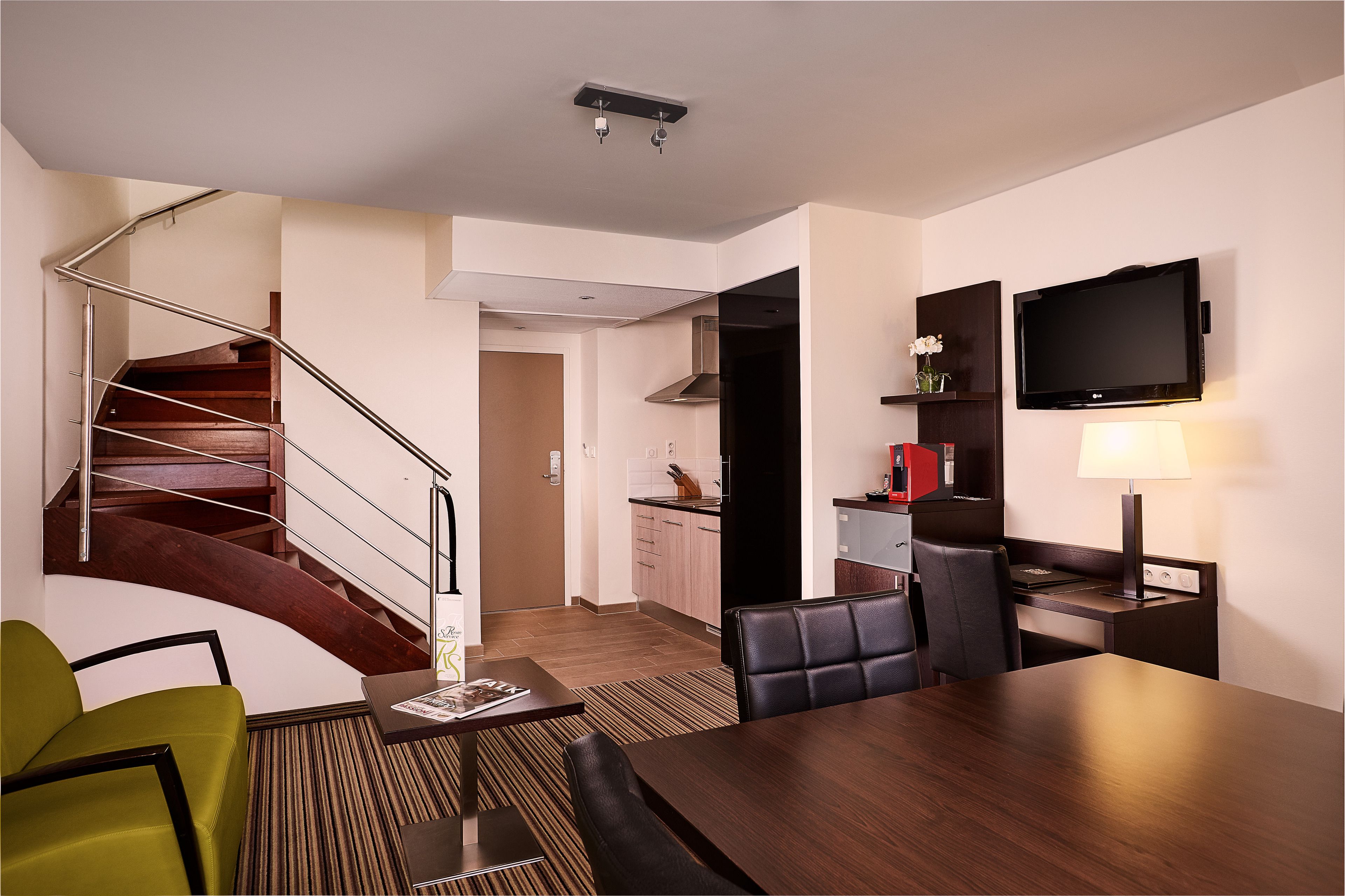 Una habitación de hotel moderna con una escalera de madera, un comedor con mesa y sillas, una cocina y un televisor de pantalla plana montado en la pared.