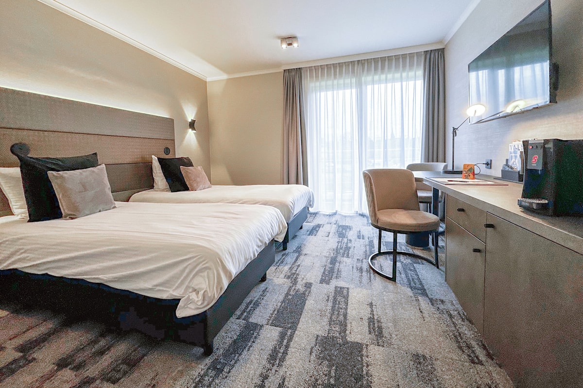 Ein Hotelzimmer mit zwei Betten, einem Schreibtisch, einem Stuhl, einem Fernseher und einem Fenster mit Vorhängen.