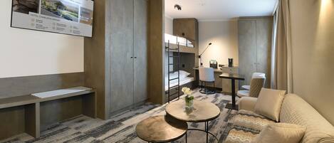 A modern hotel room with a sofa, a round table, a bench, and a wall-mounted TV displaying hotel information.