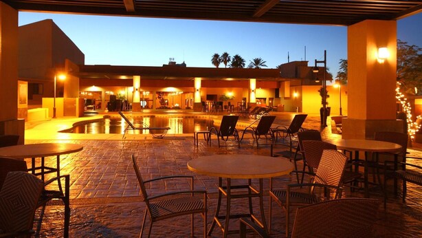 Zona de piscina al aire libre con sillones reclinables y mesas, rodeada de edificios con iluminación cálida.
