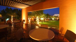Un patio al aire libre con mesas y sillas redondas, una zona de estar cubierta y vistas a un patio iluminado al anochecer.