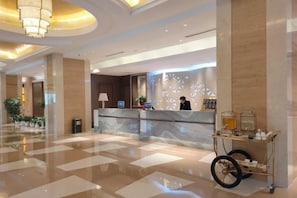Zona de recepción de hotel con mostrador de mármol, pantalla digital y carrito de bebidas.