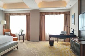 Una habitación de hotel con cama, escritorio, silla, televisión y ventanas grandes con cortinas.