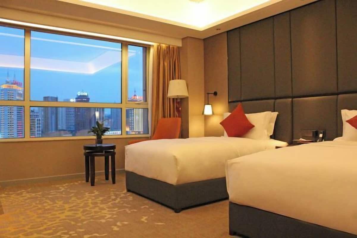 A hotel room with two beds, a large window with a city view, a small table with a plant, and a headboard with a lamp.