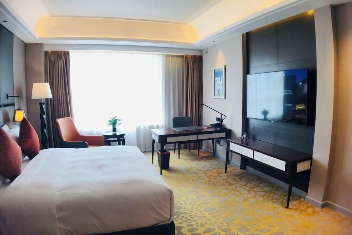 Una habitación de hotel con una cama grande, un escritorio con silla, televisión y ventana con cortinas.