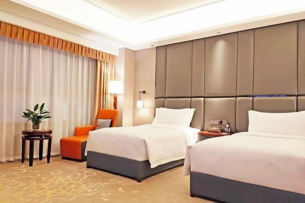 A hotel room with two beds, a small table with a plant, an orange chair, and a large padded headboard.