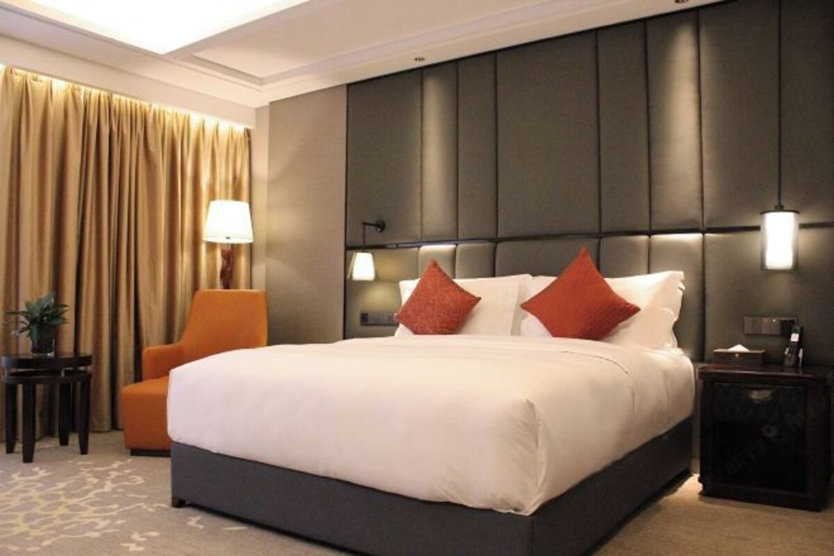 A hotel room with a large bed, a headboard with built-in lighting, a side table with a lamp, and a chair.