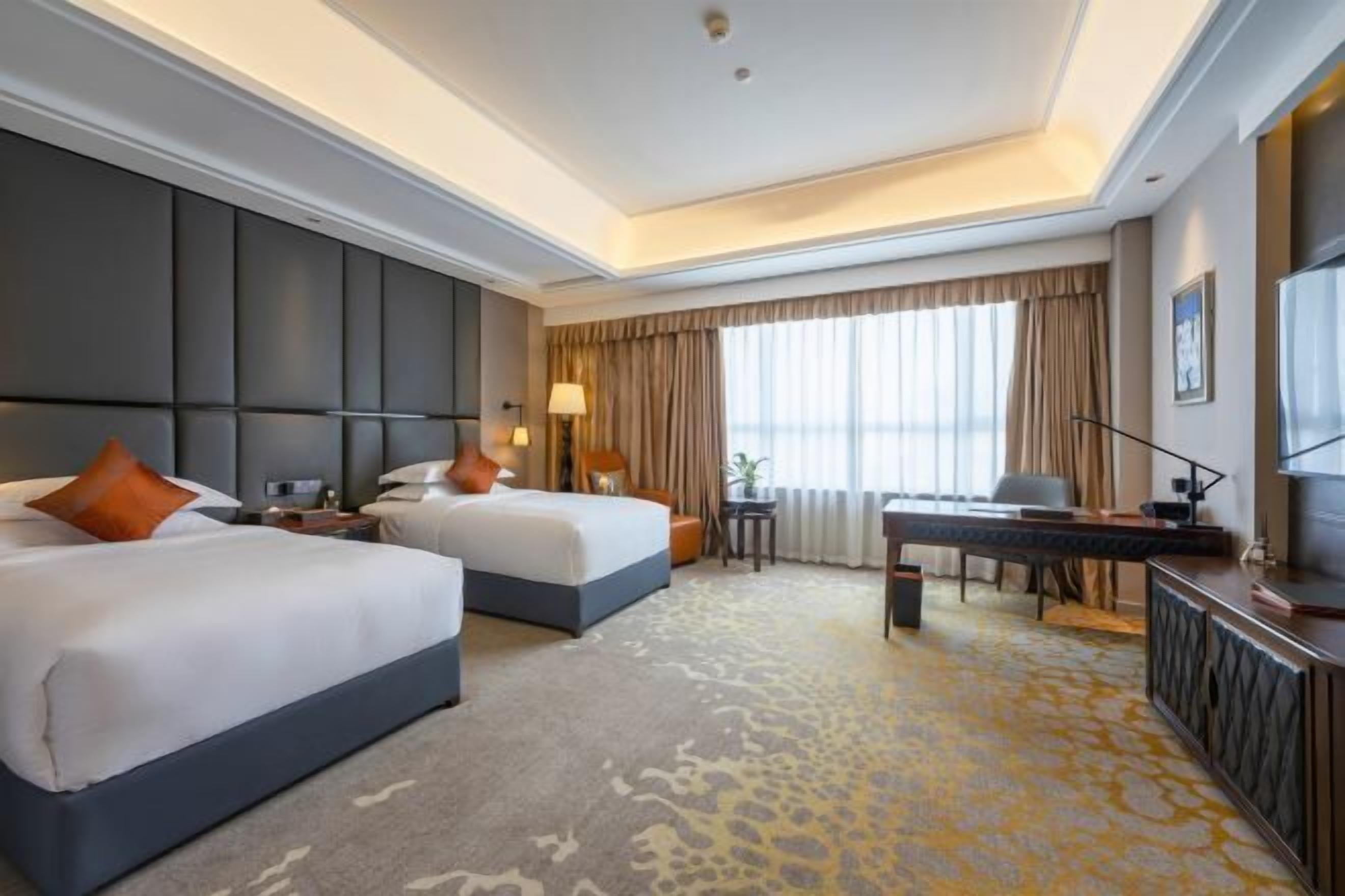 Quarto de hotel com duas camas, uma mesa de trabalho e uma janela grande com cortinas.