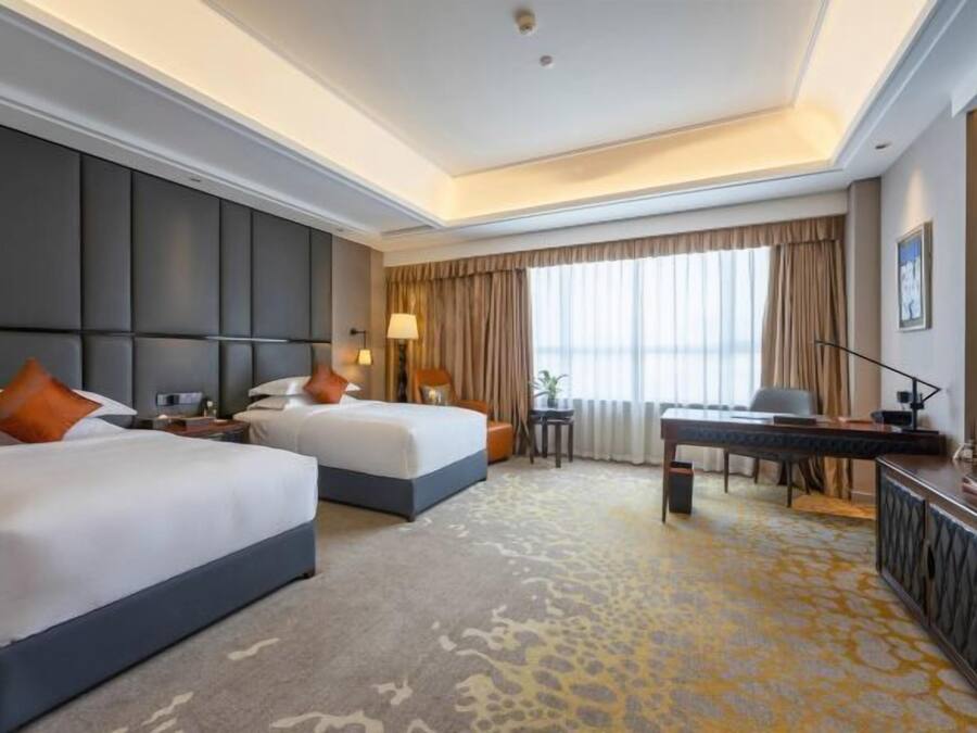 Quarto de hotel com duas camas, uma mesa de trabalho e uma janela grande com cortinas.