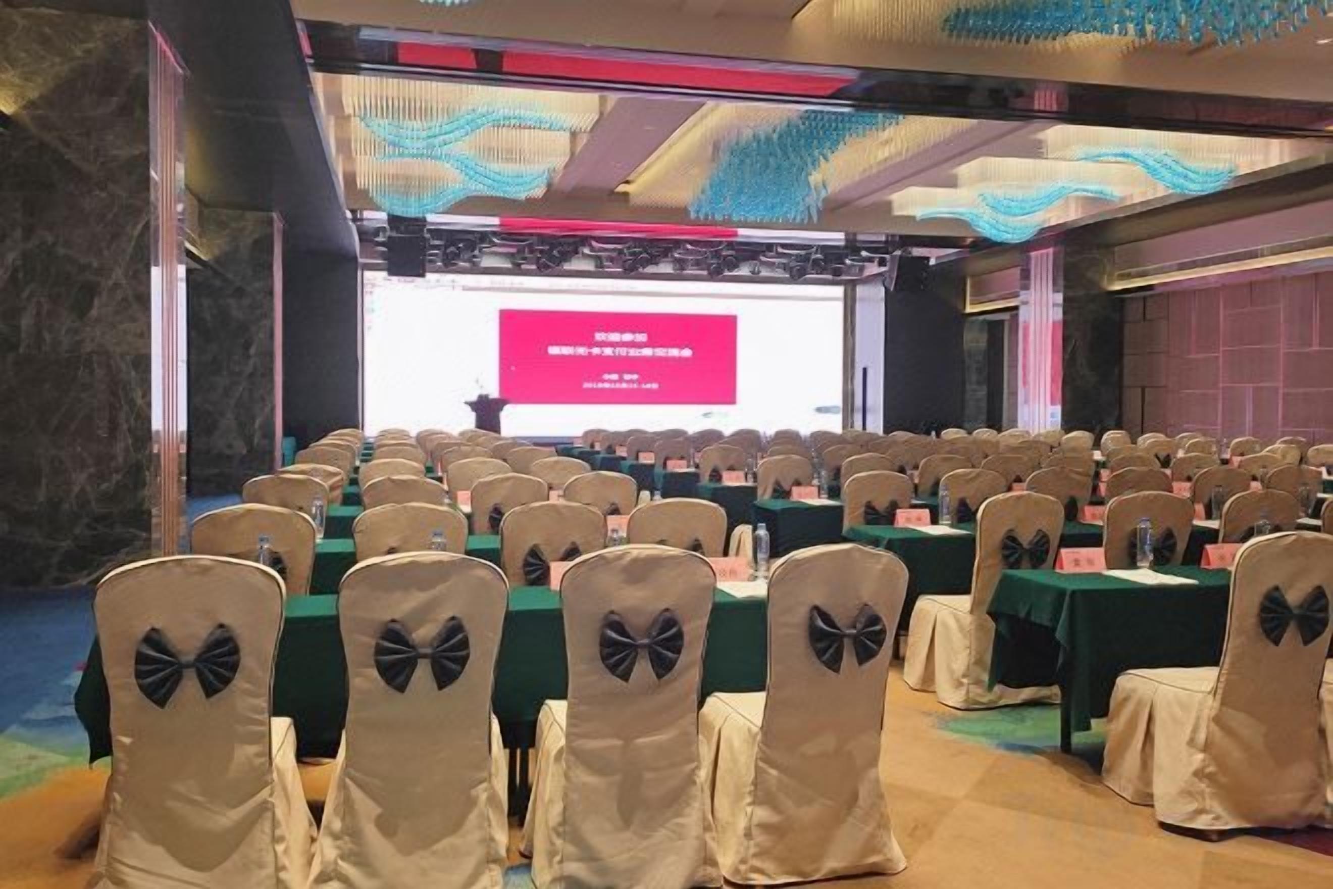 Une salle de conférence avec des rangées de tables et de chaises, une scène dotée d’un écran de présentation et un plafond décoratif.