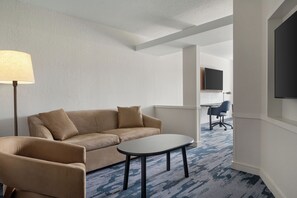 A hotel room with a sofa, a small table, a lamp, a TV, and a desk with a chair.