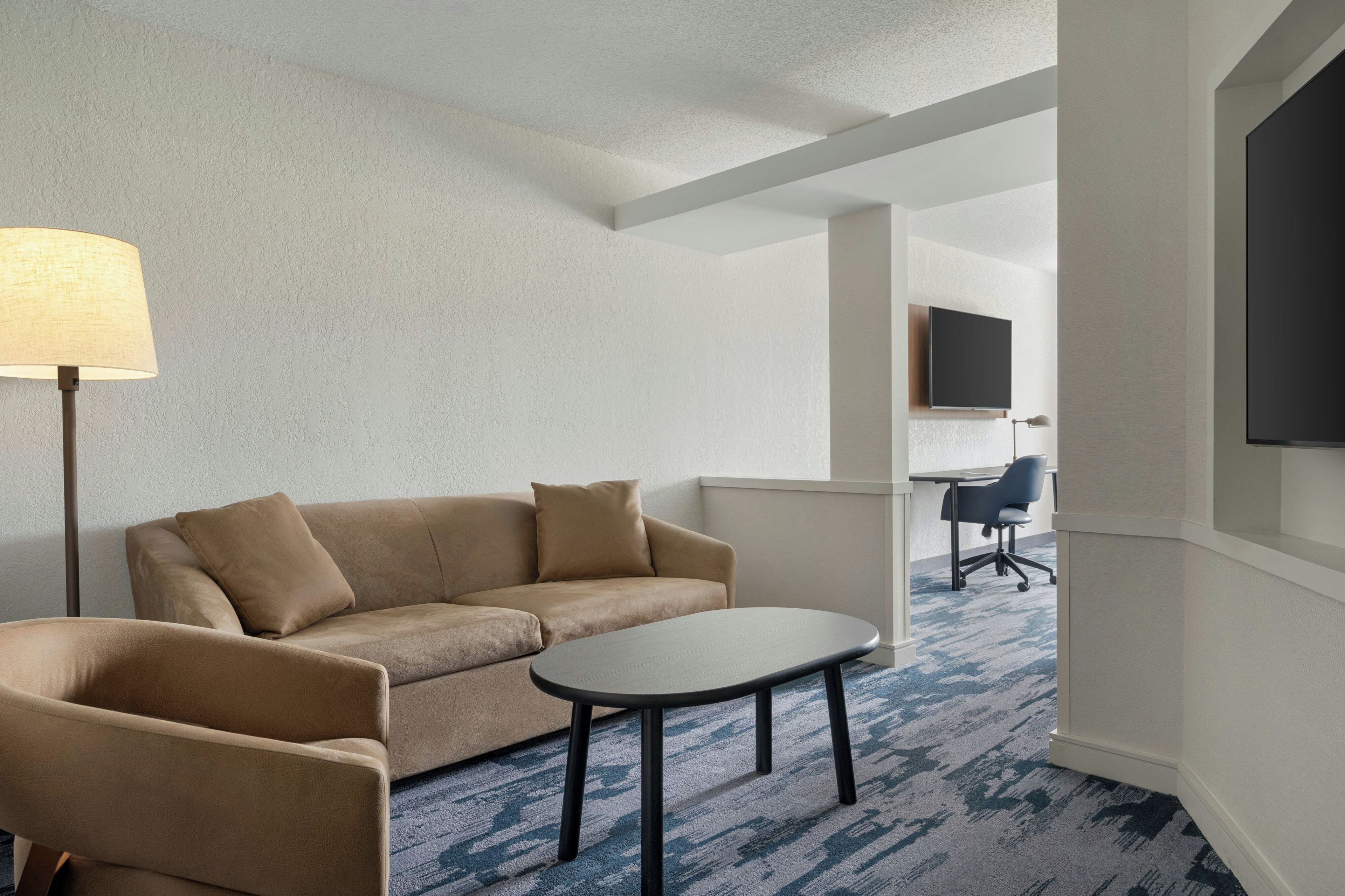 A hotel room with a sofa, a small table, a lamp, a TV, and a desk with a chair.