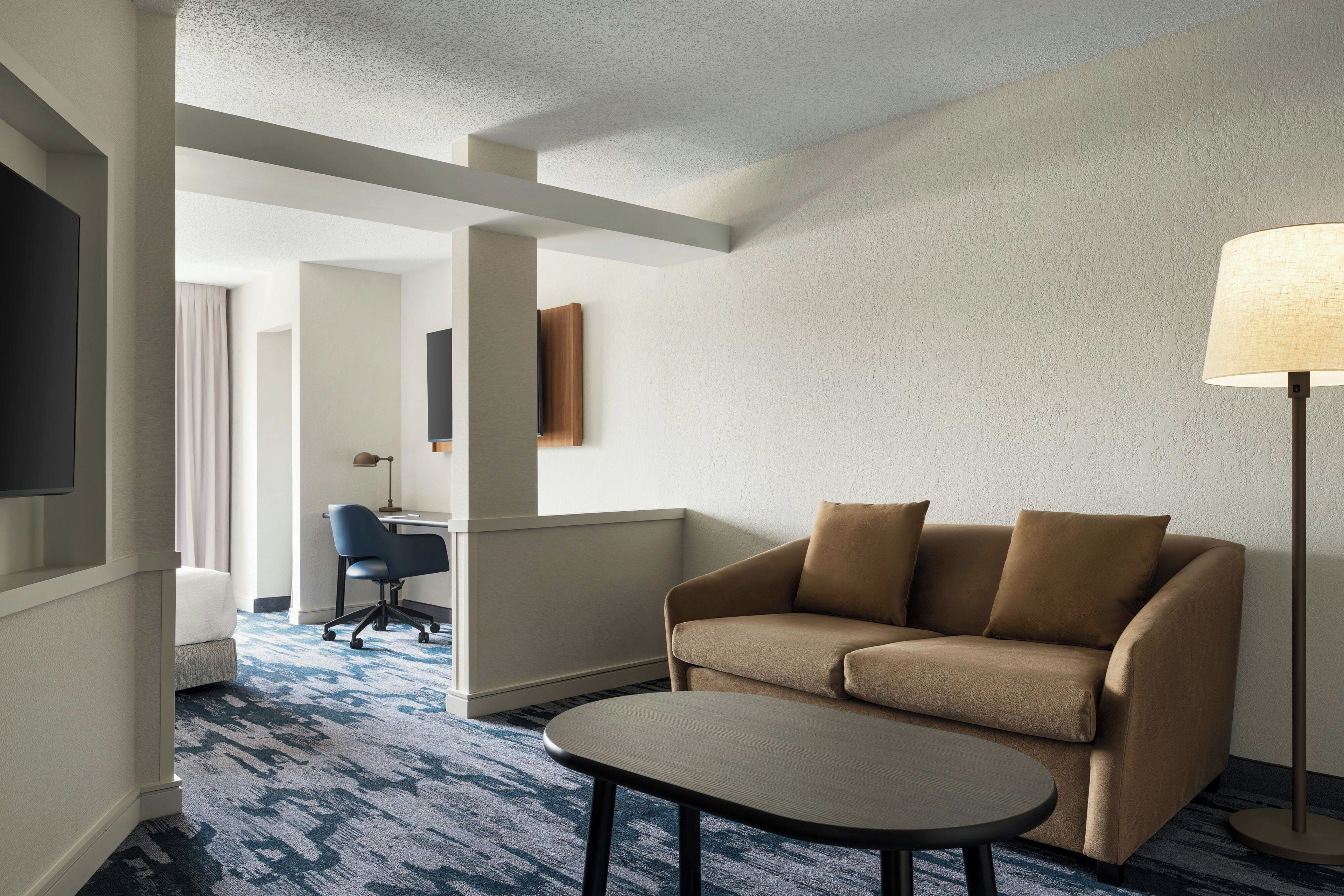 A hotel room with a sofa, a small table, a lamp, a TV, and a desk with a chair.