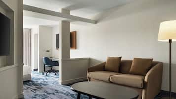 A hotel room with a sofa, a small table, a lamp, a TV, and a desk with a chair.