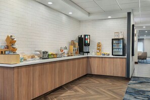 Una zona de recepción de hotel con un mostrador de madera, dispensador de bebidas y un refrigerador.