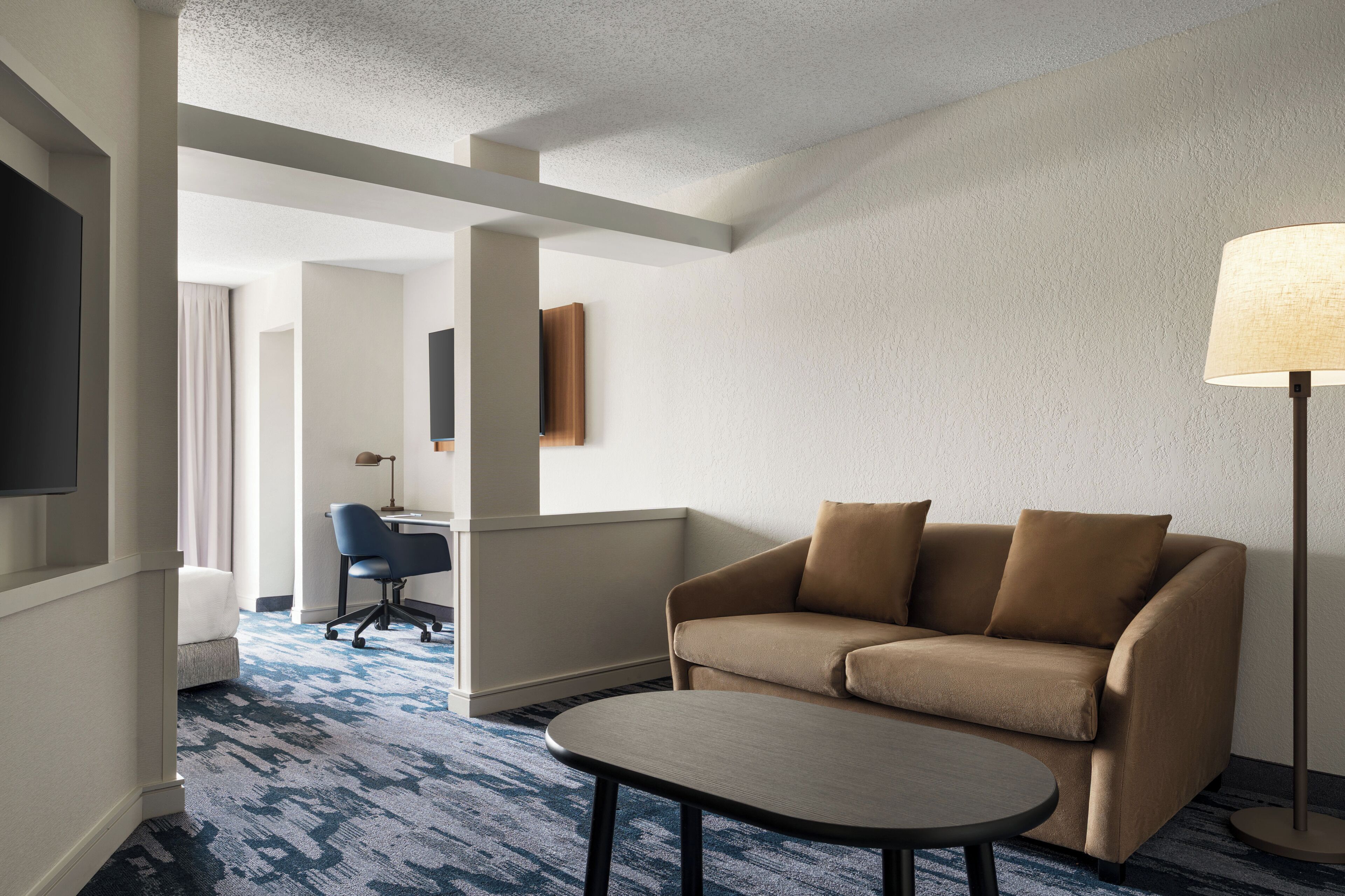 A hotel room with a sofa, a small table, a lamp, a TV, and a desk with a chair.