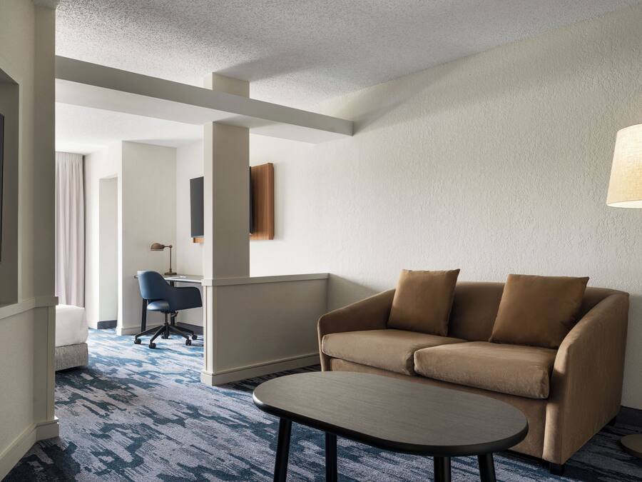 A hotel room with a sofa, a small table, a lamp, a TV, and a desk with a chair.