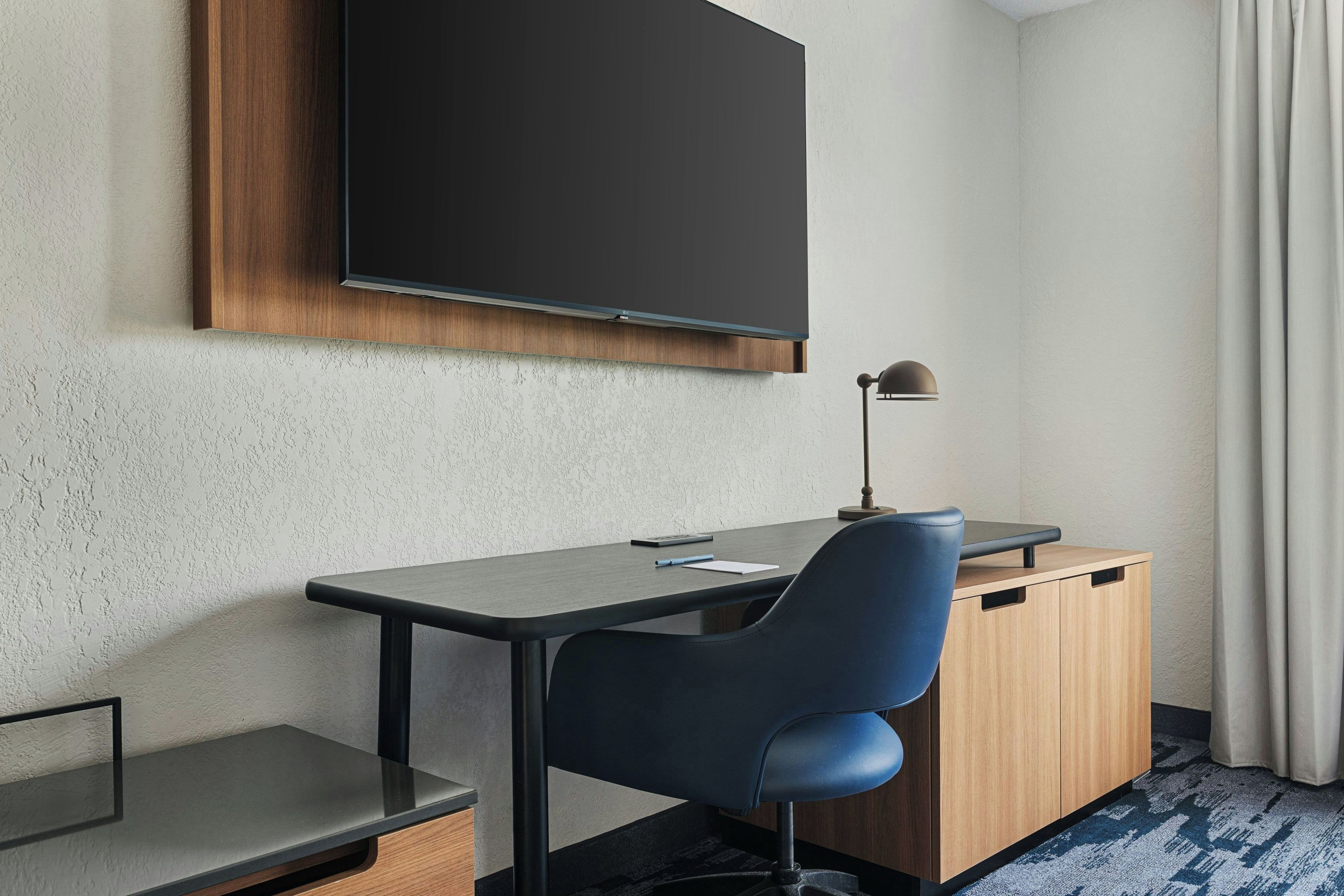 A hotel room with a desk, chair, television, and a lamp.