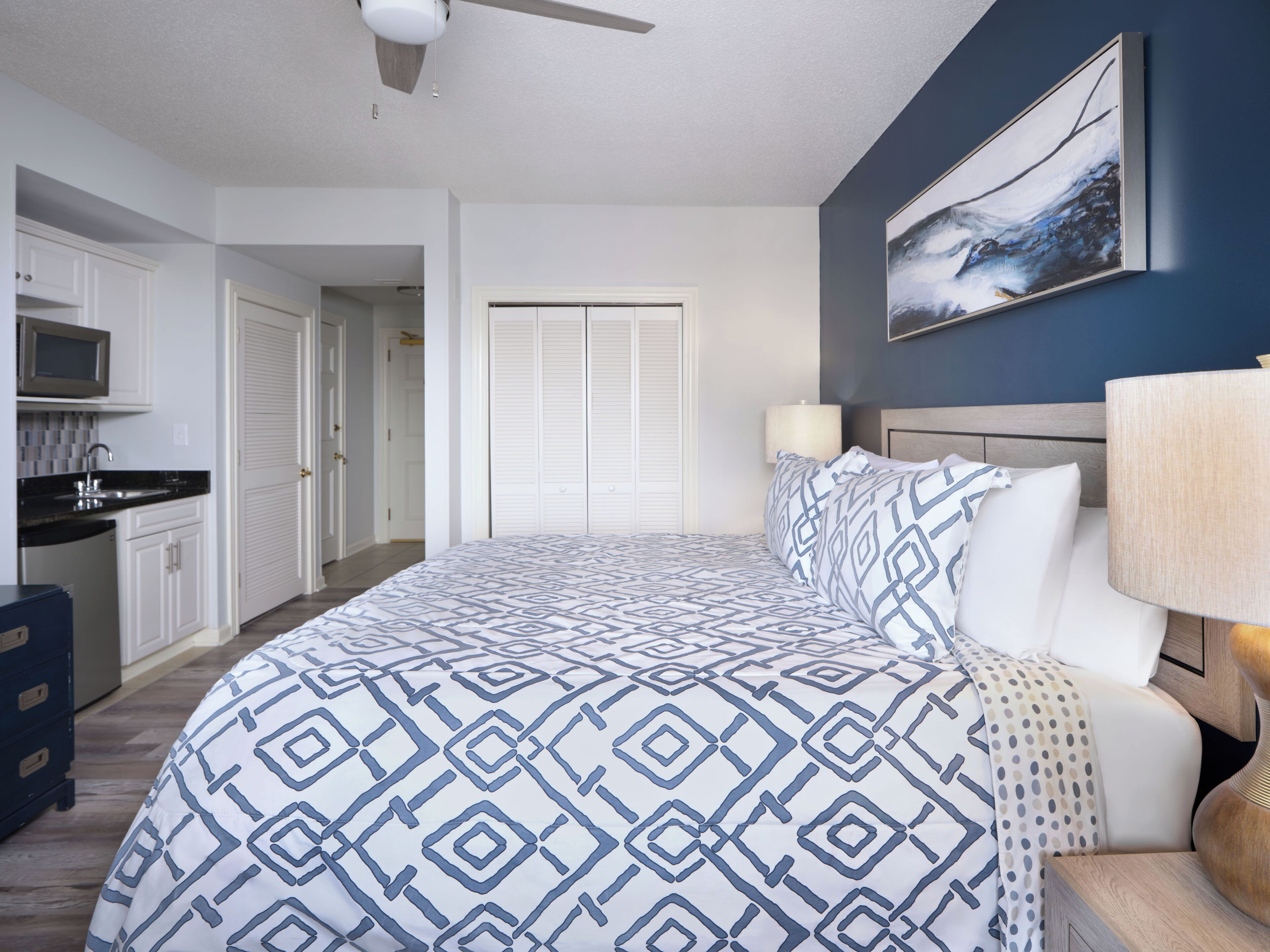 A bedroom with a bed, a nightstand, a lamp, a painting on the wall, a ceiling fan, and a view of the kitchen and bathroom.
