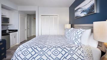 A bedroom with a bed, a nightstand, a lamp, a painting on the wall, a ceiling fan, and a view of the kitchen and bathroom.