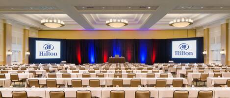 Un salón de conferencias amplio con filas de mesas y sillas, un escenario con un atril y una gran pantalla que muestra el logotipo de Hilton.