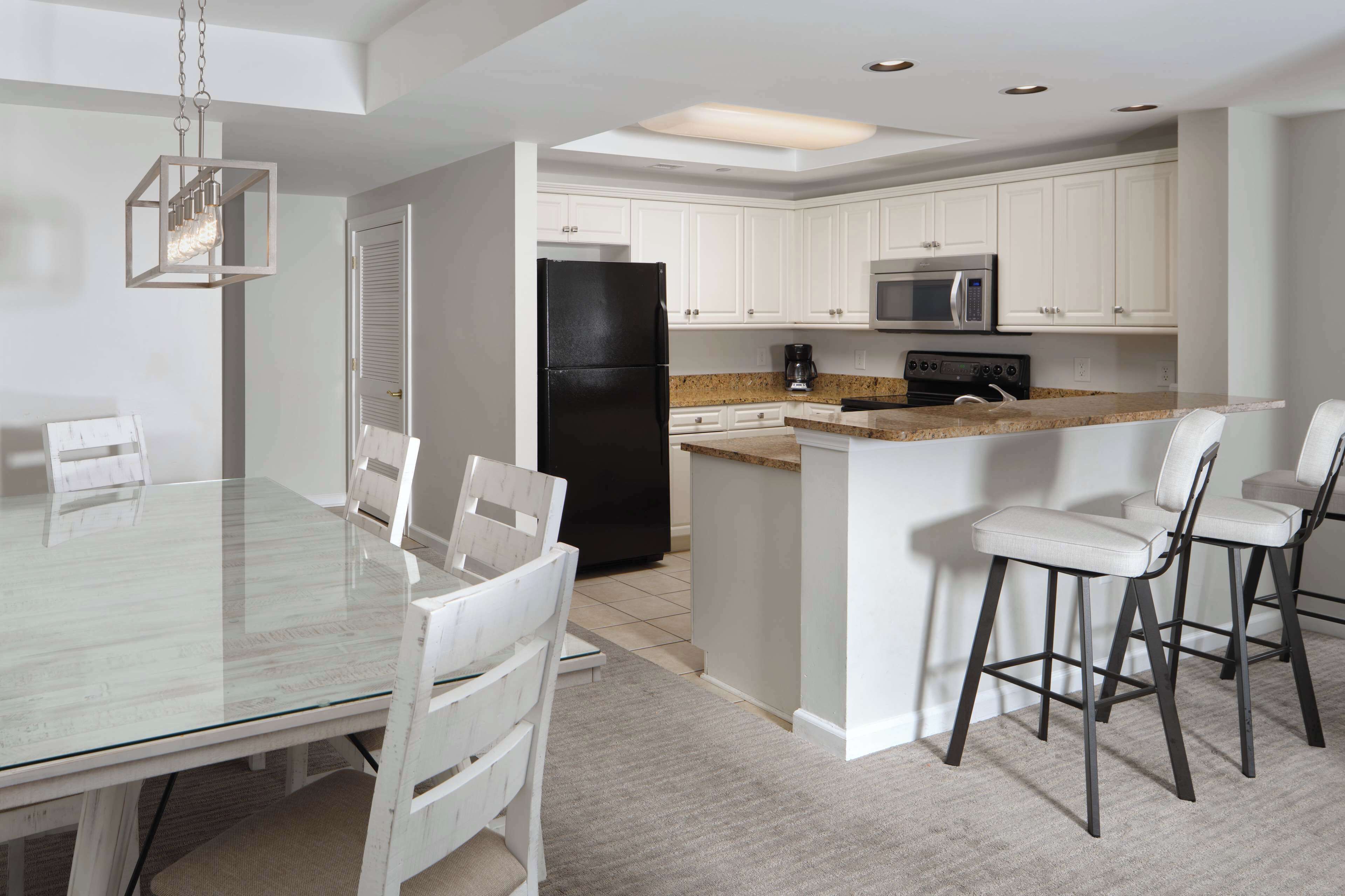 A modern kitchen with white cabinets, a black refrigerator, a microwave, and a granite countertop. There is a glass dining table with white chairs and a hanging light fixture.