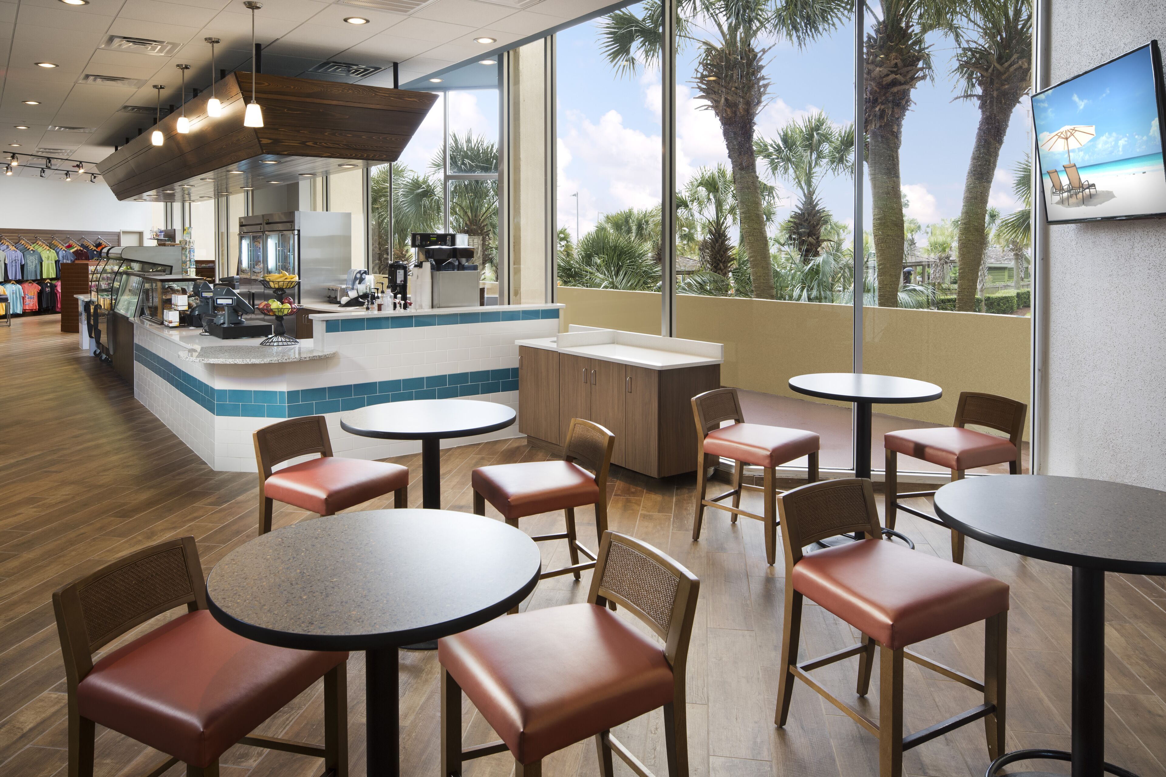 A restaurant with round tables and chairs, a counter with a display case, and a view of palm trees through large windows.