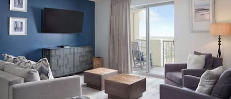 A modern living room with a flat-screen TV, grey sofas, wooden ottomans, and a balcony with a view.