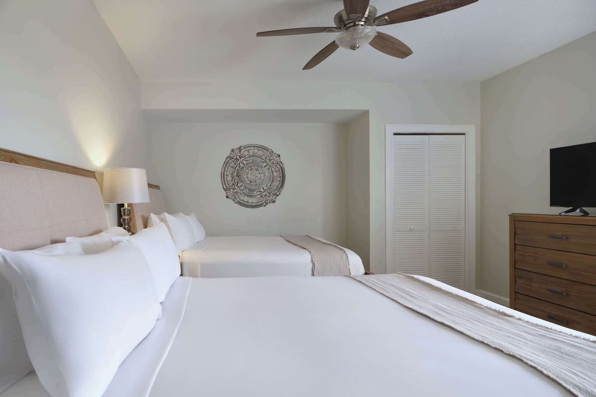 Une chambre d’hôtel avec deux lits, un ventilateur de plafond, un élément décoratif mural, une télévision et une commode en bois.