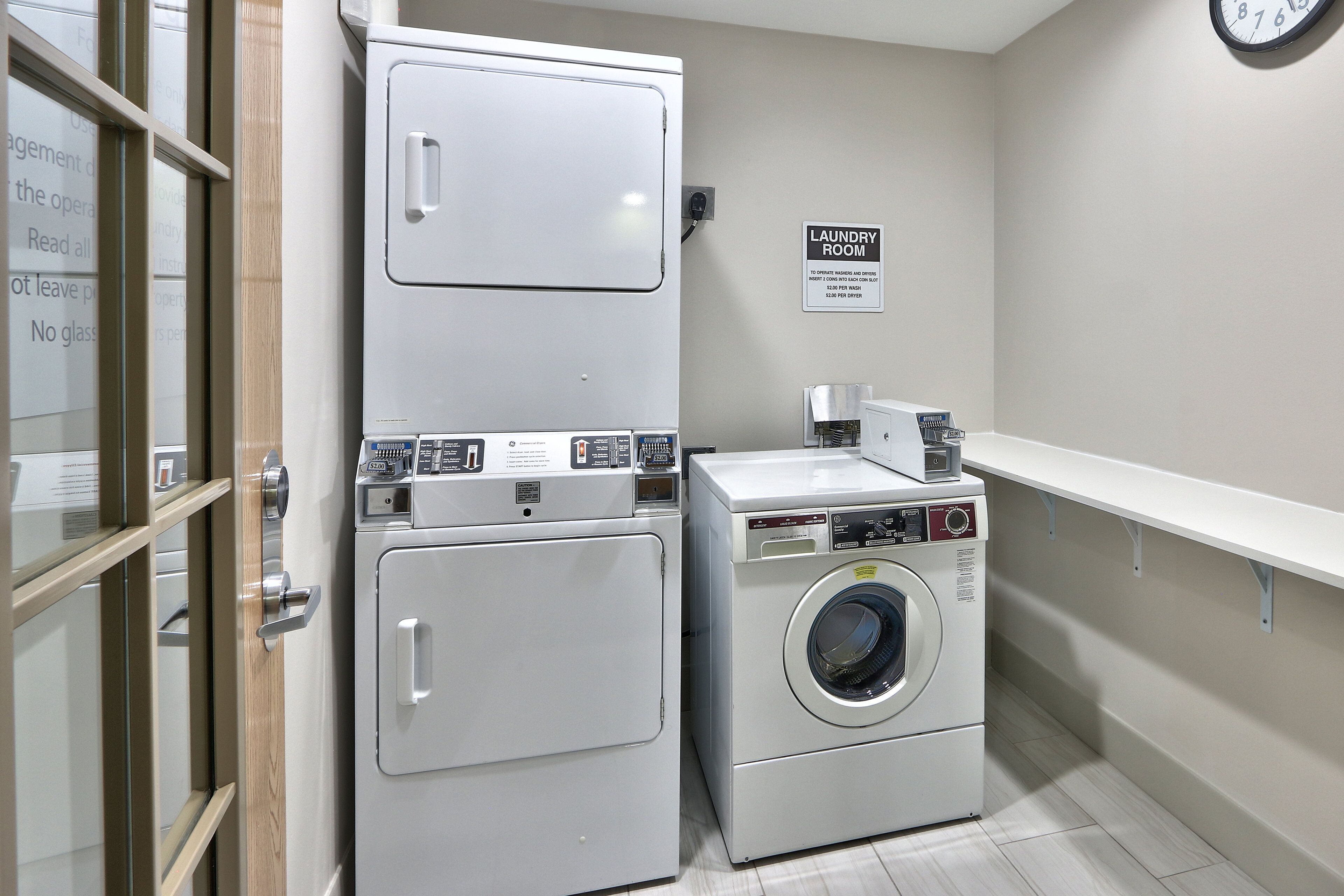 A laundry room with two stacked washers and a front-loading dryer, a clock on the wall, and a sign indicating the room's purpose.