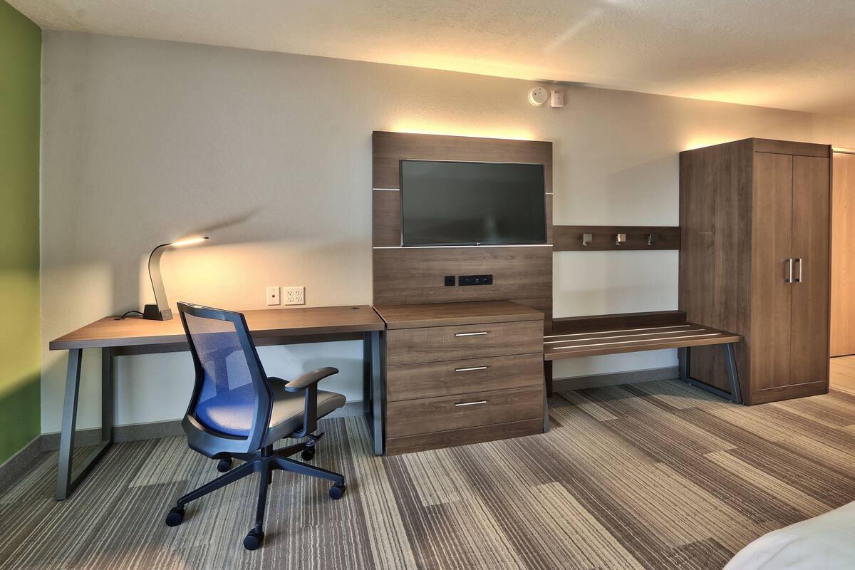 Ein Hotelzimmer mit einem Holztisch, einem Sessel, einem an der Wand montierten Fernseher und einem Kleiderschrank.