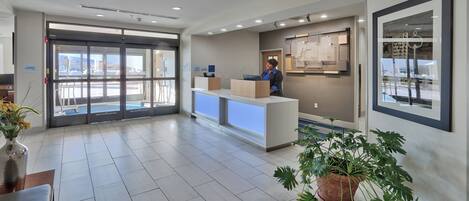 A hotel reception area with a reception desk, a large potted plant, and a view of the outside through the glass doors.