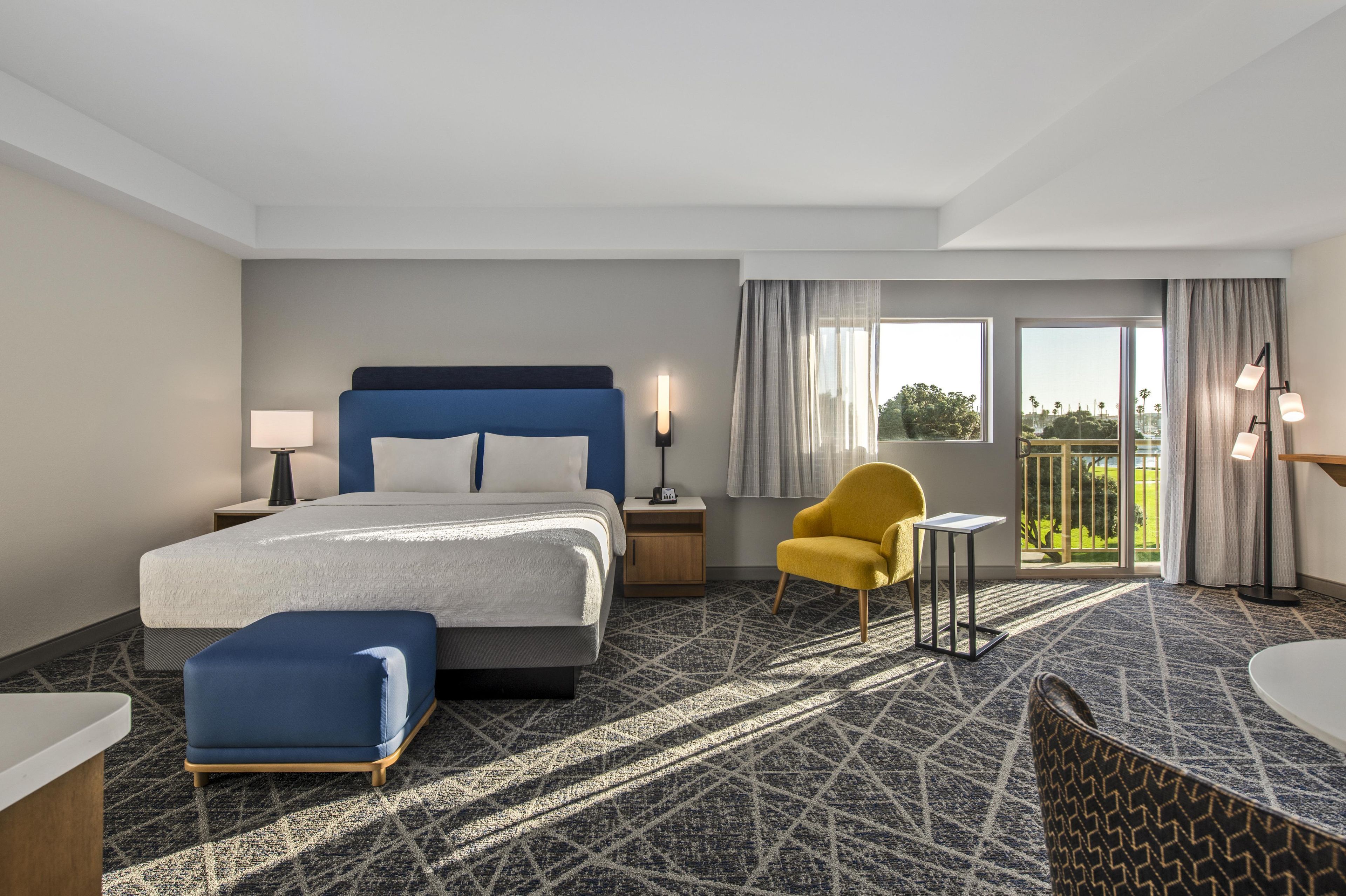 Ein Hotelzimmer mit Bett, Stuhl, kleinem Tisch, Nachttisch und Blick ins Freie.