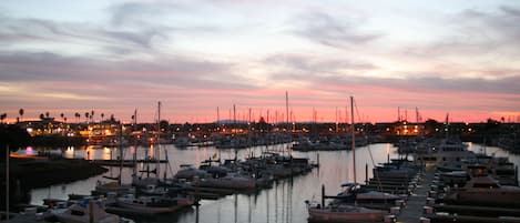 Un puerto deportivo al atardecer con barcos amarrados, agua tranquila y una costa lejana con edificios.