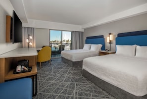 A hotel room with two beds, a desk, a chair, a TV, and a balcony with a view of boats.