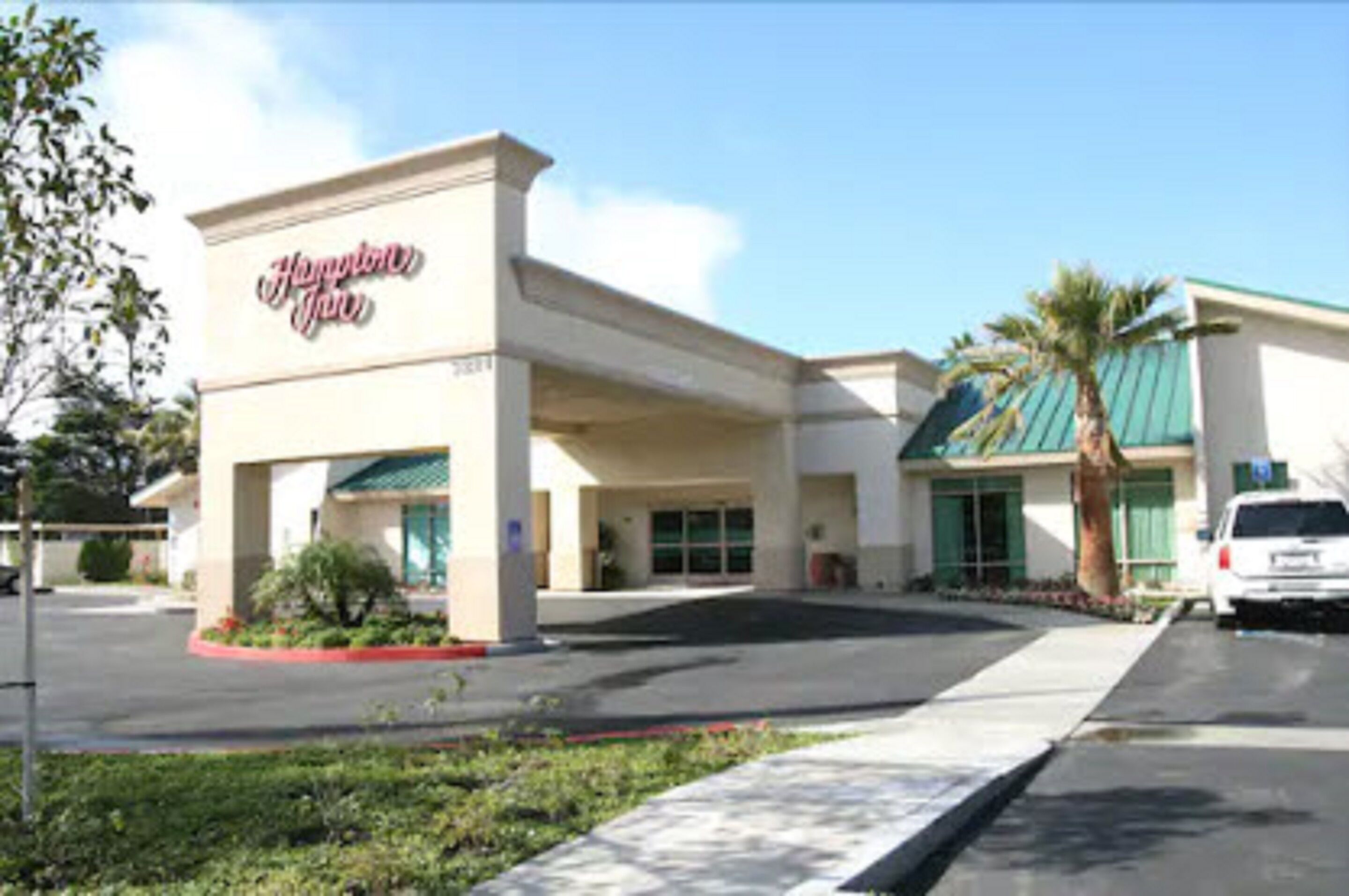 Un hotel Hampton Inn con fachada moderna, un área de estacionamiento techada y una palmera en primer plano.